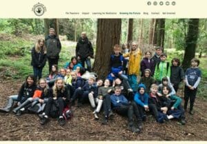 A group of children and adults pose for a photo in a forest, surrounded by tall trees and greenery, with some sitting and others standing.